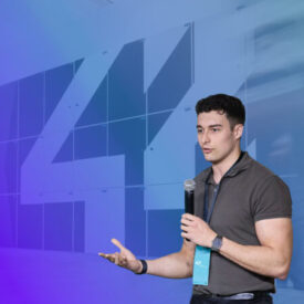 Ponente joven con camisa polo gris dando una presentación en el Campus 42 Madrid, sosteniendo un micrófono y gesticulando, con el logotipo del número 42 en azul de fondo y llevando una pulsera azul identificativa del evento
