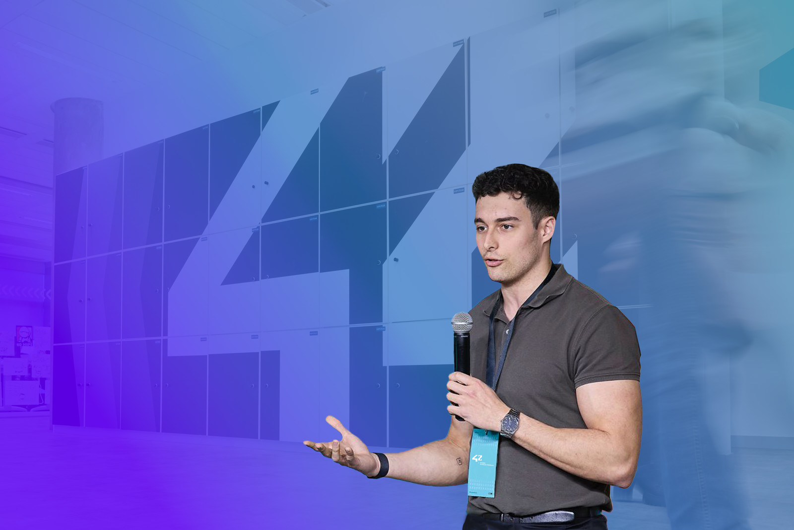 Ponente joven con camisa polo gris dando una presentación en el Campus 42 Madrid, sosteniendo un micrófono y gesticulando, con el logotipo del número 42 en azul de fondo y llevando una pulsera azul identificativa del evento