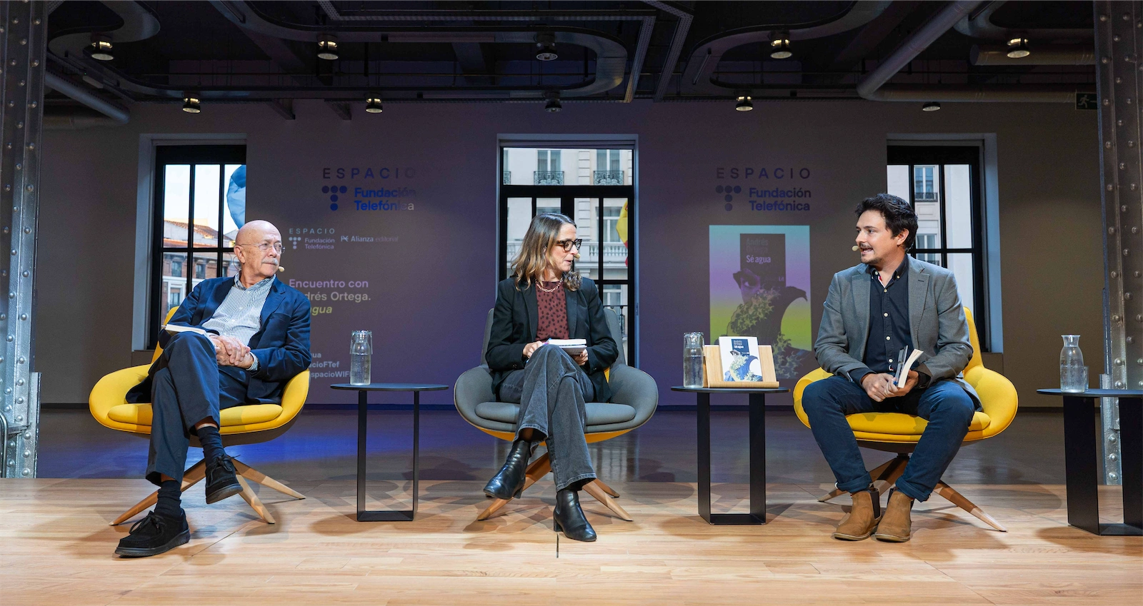 Andrés Ortega, Carola García-Calvo y Jorge Morla están sentados en el escenario durante un evento de la Fundación Telefónica. Los tres están en sillas amarillas mientras conversan, y se ven detalles del público en el fondo, escuchando atentamente. La pantalla detrás muestra el cartel del evento con el título 
