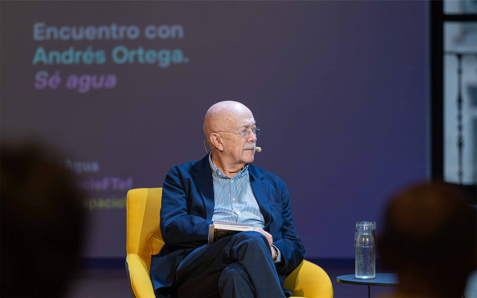 Andrés Ortega está sentado en una silla amarilla, mirando hacia un lado durante un evento en el que aparece el texto 