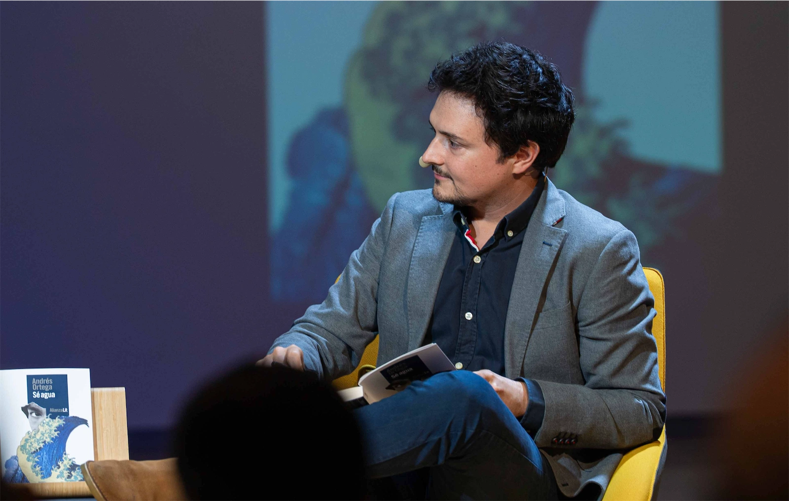 Jorge Morla, con cabello oscuro y corto, está sentado en una silla amarilla, mirando hacia la cámara. Sostiene un libro en una charla sobre el libro 