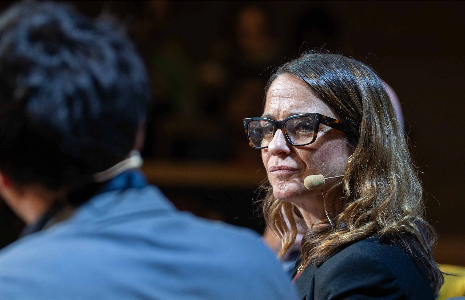 Carola García-Calvo, con gafas grandes y cabello castaño largo, mira fijamente mientras escucha a otra persona durante una charla.