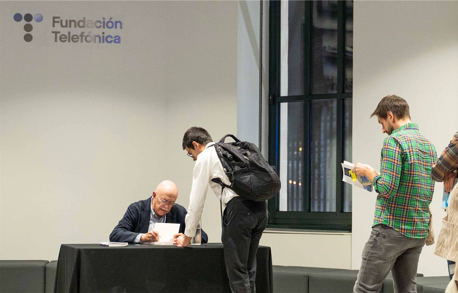 Andrés Ortega firma un libro para un joven que lleva una mochila, en un evento organizado por la Fundación Telefónica. El fondo muestra el logo de la fundación en una pared blanca.