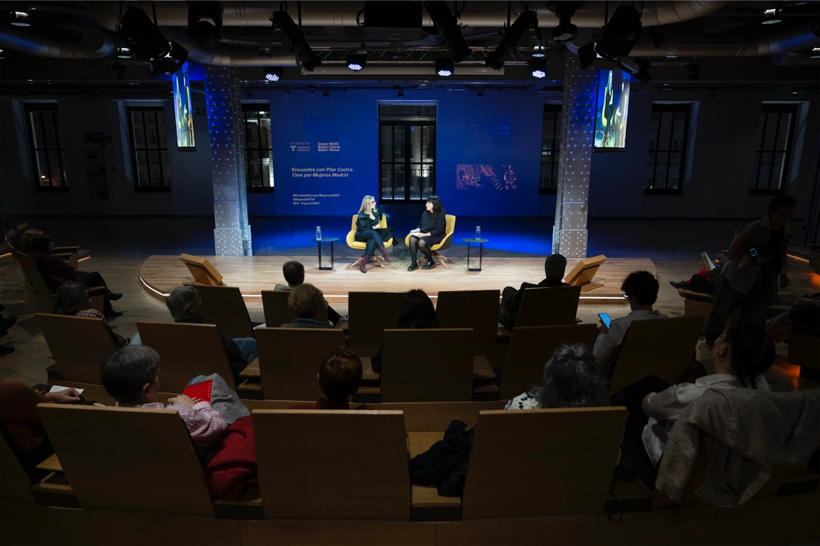 Vista general del evento con Pilar Castro y María Adell sentadas en sillones amarillos mientras la audiencia observa, en un espacio iluminado de manera moderna.
