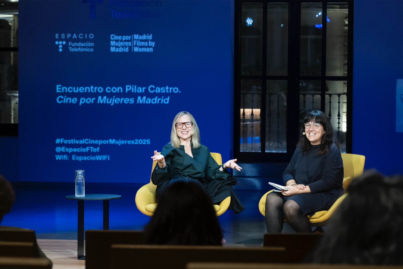 Pilar Castro gesticulando mientras habla y María Adell sonriendo mientras la escucha, ambas sentadas en sillones amarillos durante una charla en un evento.