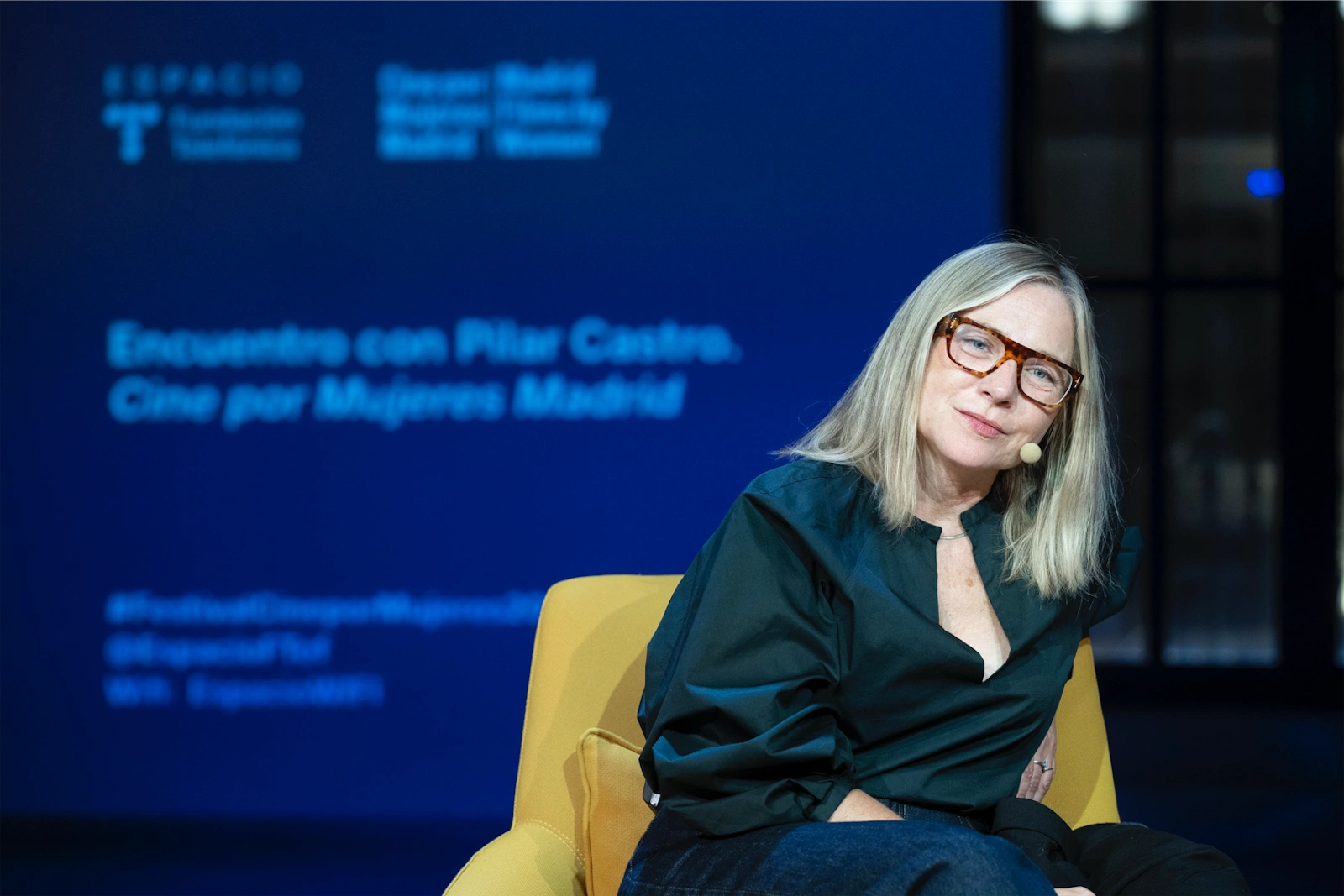 Pilar Castro sentada en un sillón amarillo durante un evento, mirando hacia la cámara con una expresión relajada mientras lleva gafas y una blusa oscura