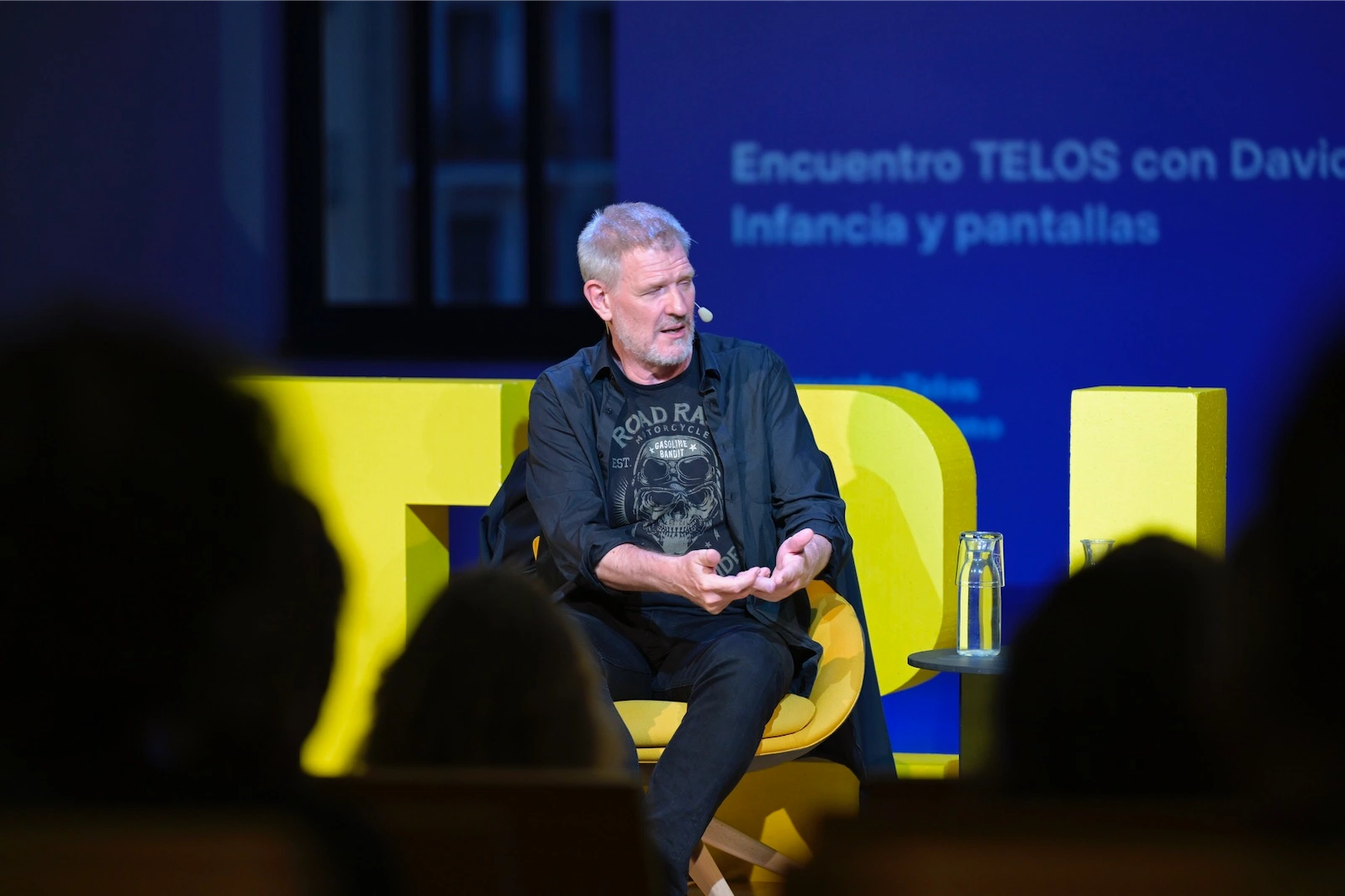 Primer plano de David Bueno hablando al público durante el Encuentro TELOS. Está sentado con las piernas cruzadas, gesticulando con las manos y usando micrófono de diadema, con fondo azul proyectado con el título del evento.