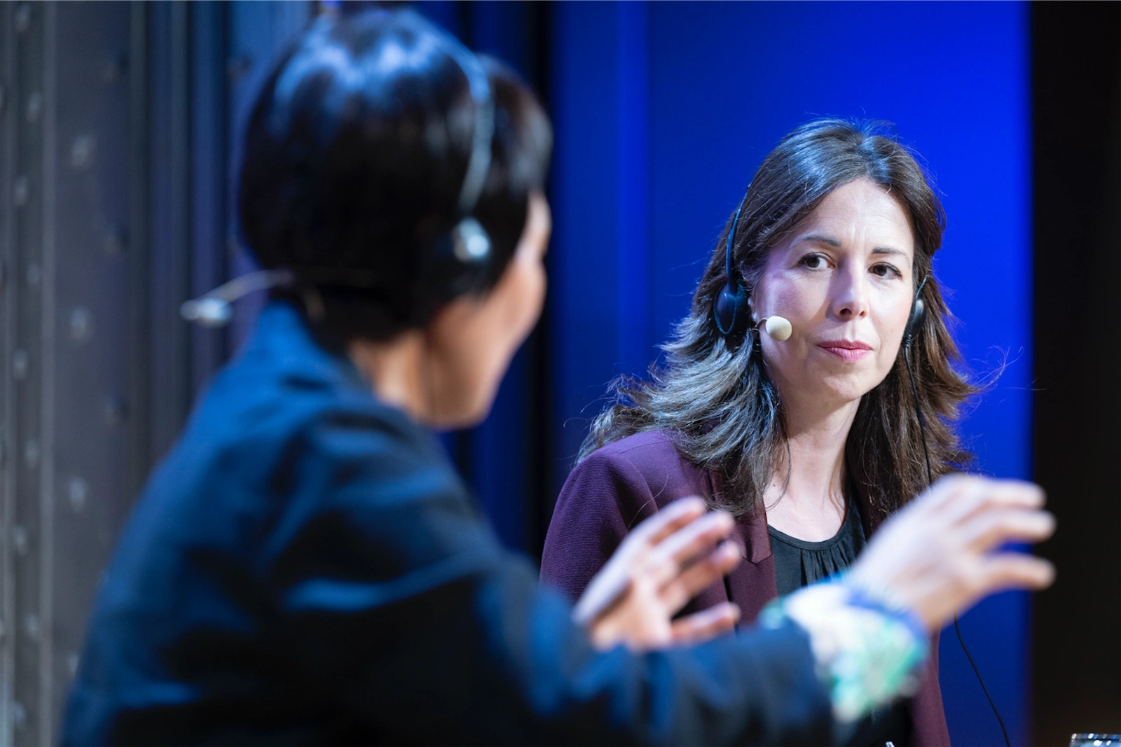 Keum Suk Gendry-Kim (izquierda) escucha atentamente durante una entrevista. Está usando auriculares con micrófono, mientras Sara Romero, que se encuentra en el fondo de la imagen, se muestra enfocada en la conversación.