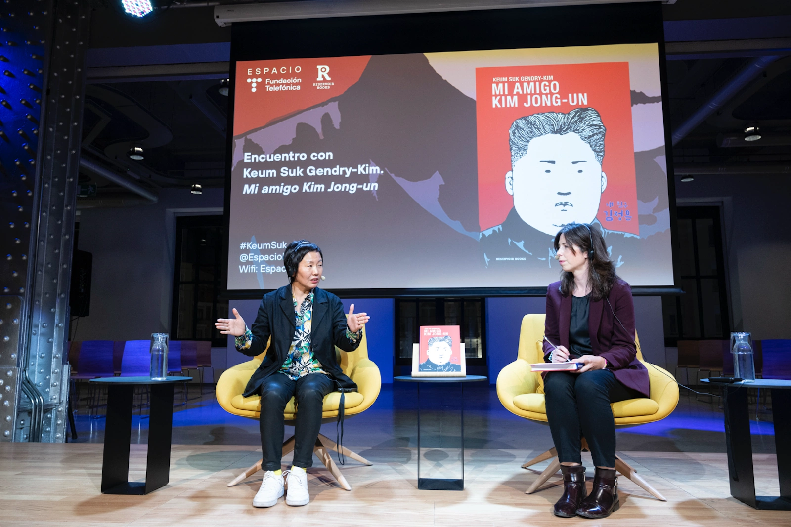 En el escenario, Keum Suk Gendry-Kim, vestida con una camisa estampada y un saco negro, está siendo entrevistada por Sara Romero, quien toma notas. Detrás de ellos, una pantalla muestra la portada del libro Mi amigo Kim Jong-un.