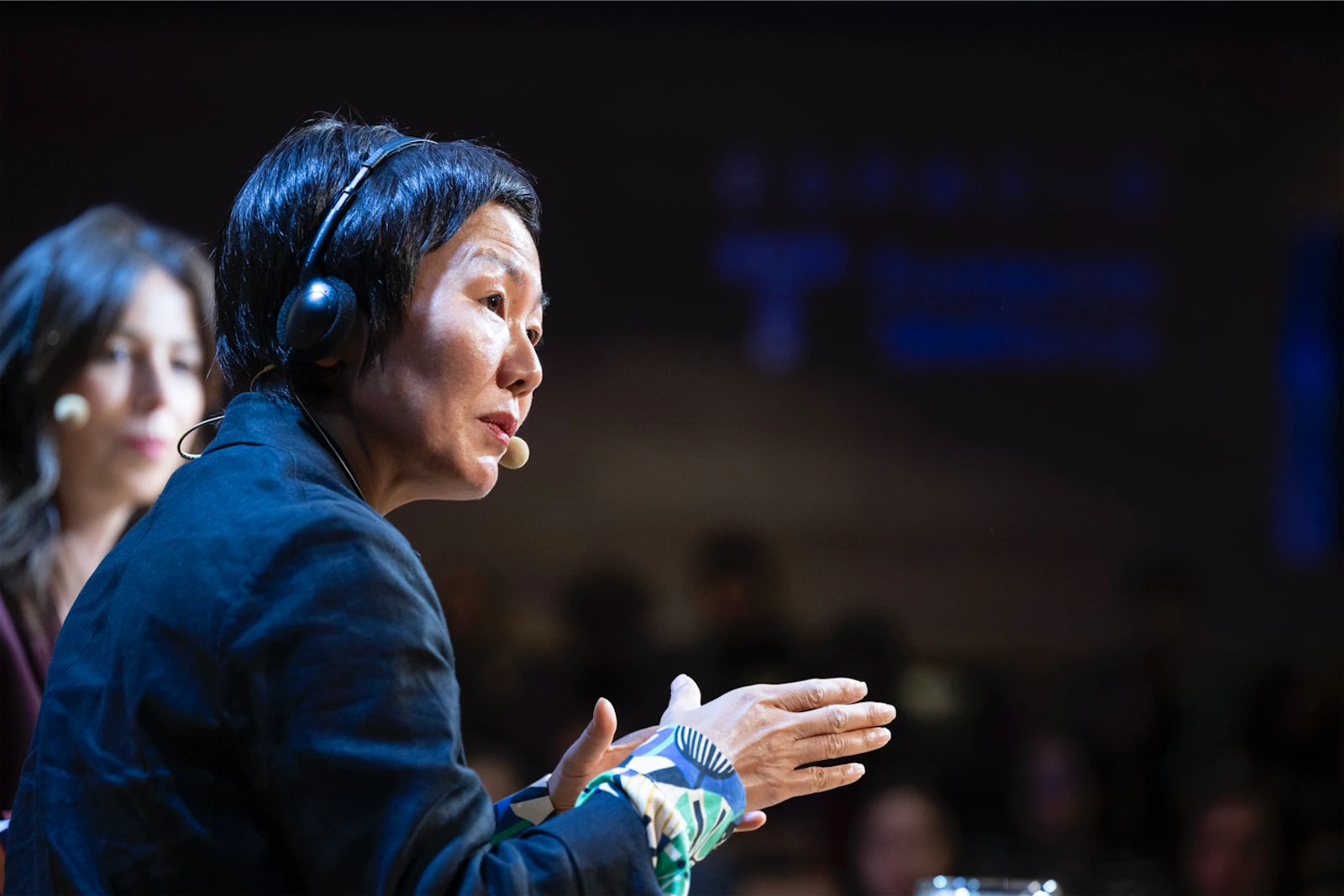 Keum Suk Gendry-Kim está hablando con entusiasmo durante la entrevista, mientras Sara Romero, en segundo plano, la observa con atención. Ambos están usando auriculares con micrófono.