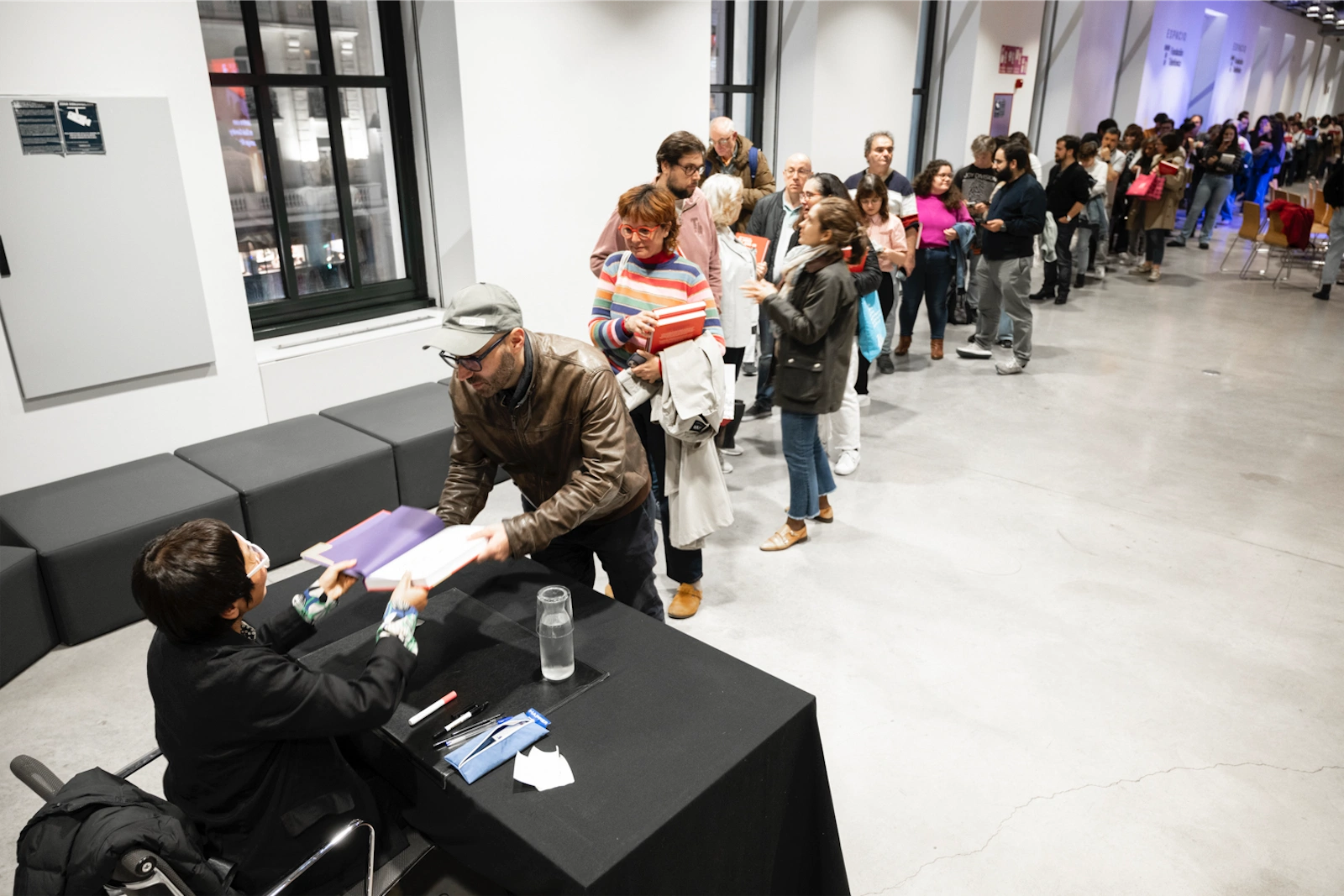 Una fila de personas espera pacientemente en un evento de firma de libros. Keum Suk Gendry-Kim está sentada a la mesa firmando libros para los asistentes.