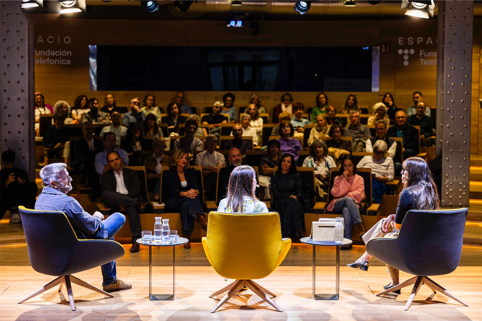 Aimar Bretos sentado en una silla amarilla con una postura relajada mientras escucha atentamente a Ángeles Caballero, quien se encuentra de espaldas en una silla gris. Al lado de Ángeles, está Paloma Rando, sentada también en una silla amarilla. El escenario está lleno de público, y la atmósfera es de una charla o evento en vivo.