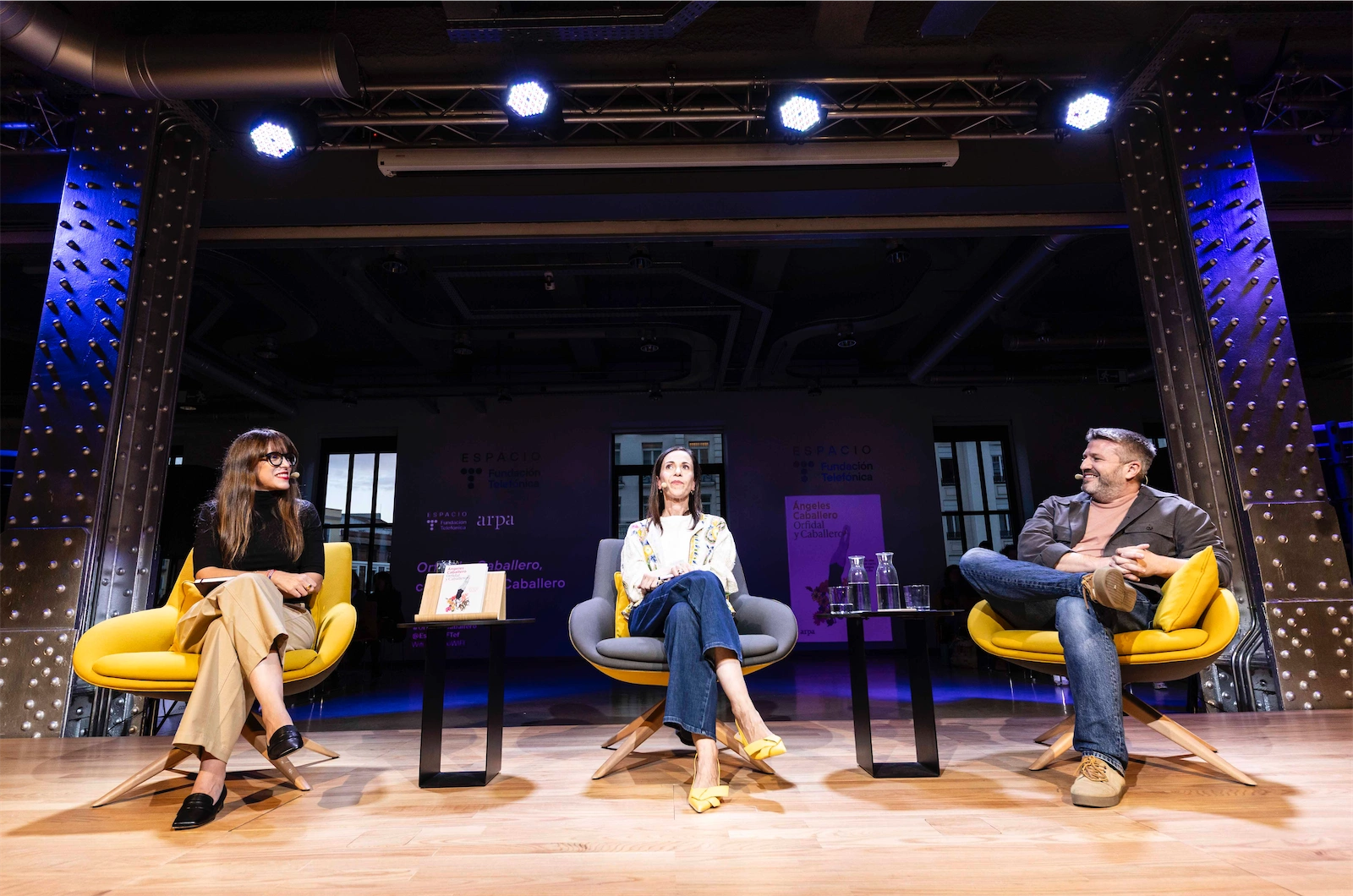 La imagen muestra a los tres (Paloma Rando, Ángeles Caballero y Aimar Bretos) sentados en el escenario. Cada uno en una silla amarilla, observando el evento o charla, con una mesa frente a ellos que tiene agua y libros, mientras el público en el fondo sigue la conversación atentamente.