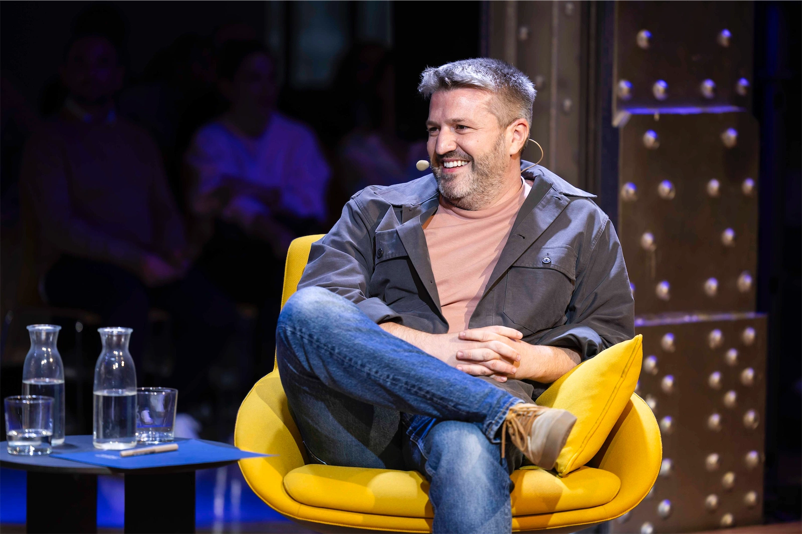 Aimar Bretos sentado con una sonrisa y actitud relajada en su silla amarilla. Lleva una camiseta color rosa claro y una chaqueta gris. Parece estar disfrutando del momento, tal vez escuchando o participando en la conversación.