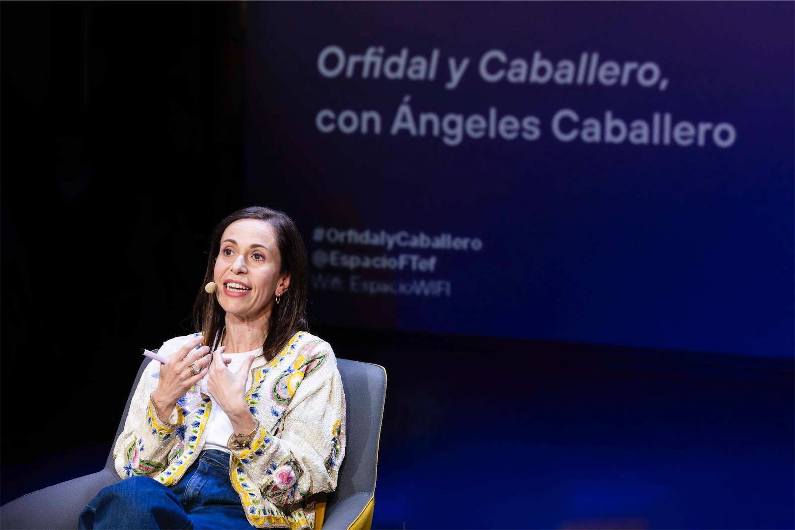 Ángeles Caballero está sentada en una silla amarilla, mirando hacia el público con una gran sonrisa mientras habla. Ella lleva una chaqueta blanca con detalles coloridos y está usando un micrófono. La pantalla detrás de ella menciona su nombre y algunos detalles sobre el evento.