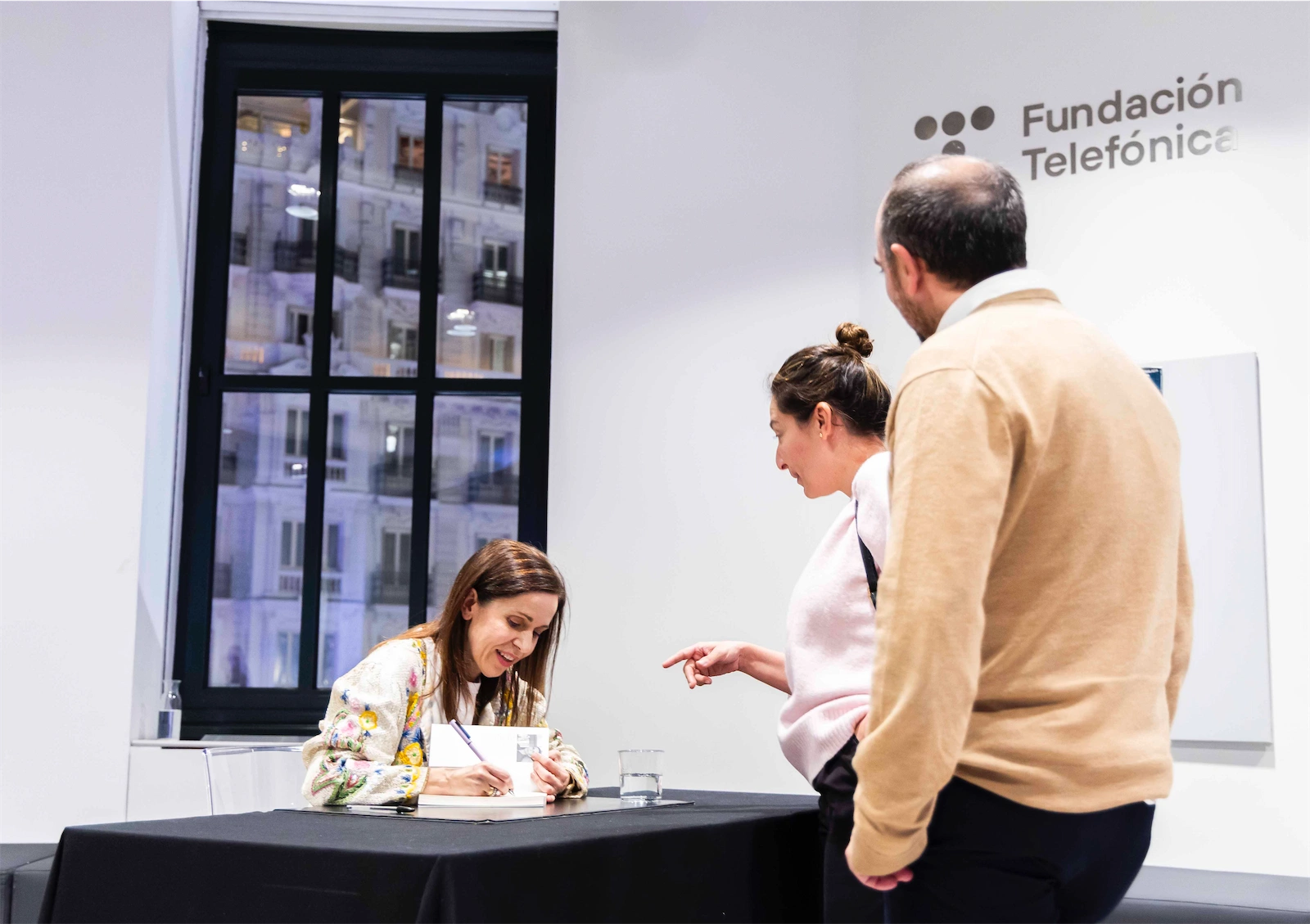 Ángeles Caballero está firmando libros para algunas personas. Se la ve sonriente y en plena interacción con los asistentes. Está en una mesa con un fondo de luz natural que entra por una ventana grande.