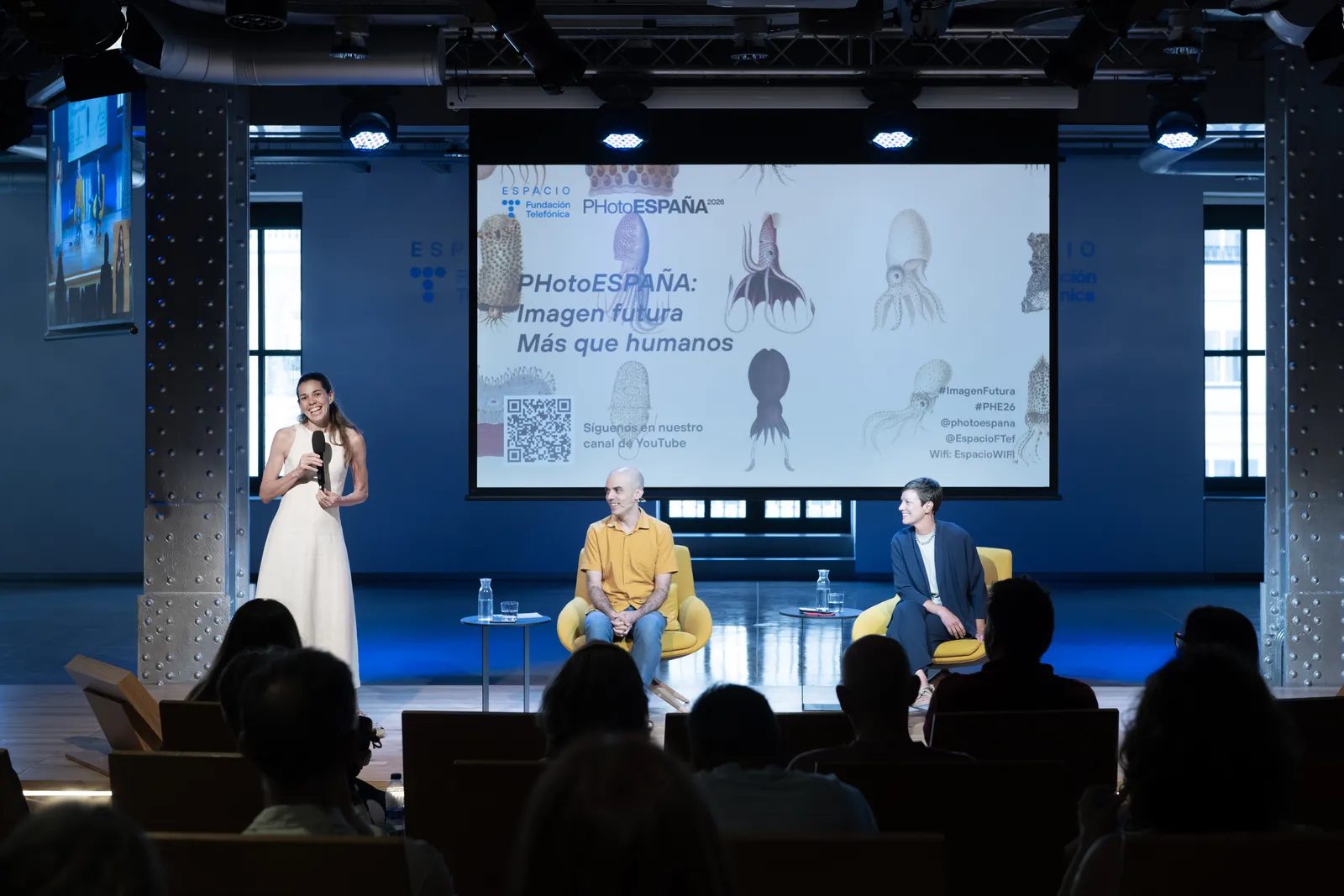 Female presenter in a white dress speaks into a microphone on stage, flanked by two male panelists in yellow and blue chairs; a large screen behind displays PHotoESPAÑA branding and marine illustrations.