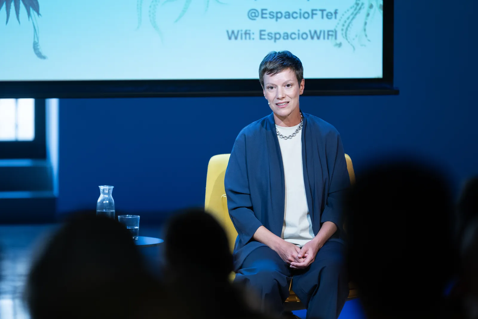 Woman speaker seated on a yellow chair on stage, blue lighting, addressing an audience with silhouettes in the foreground.