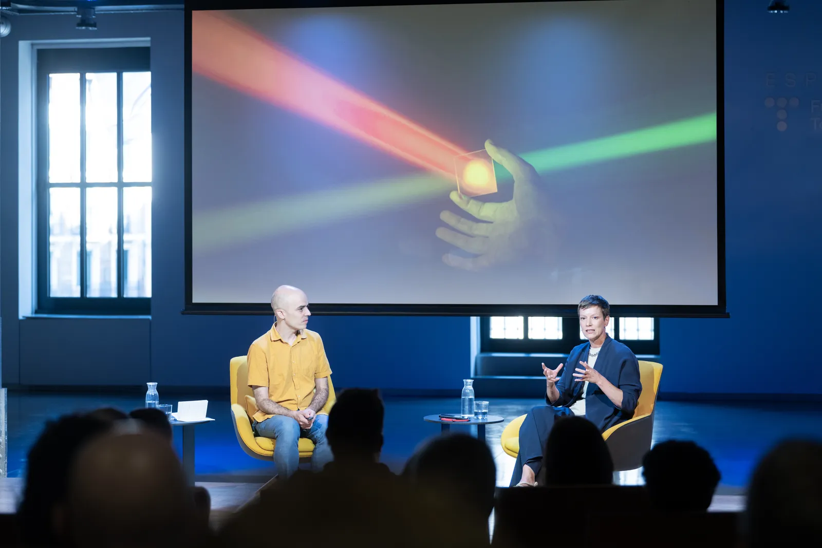 Two speakers sit in yellow chairs on a stage, with a large screen showing a glowing hand holding a light square and colorful laser beams in the background, audience silhouettes in front.