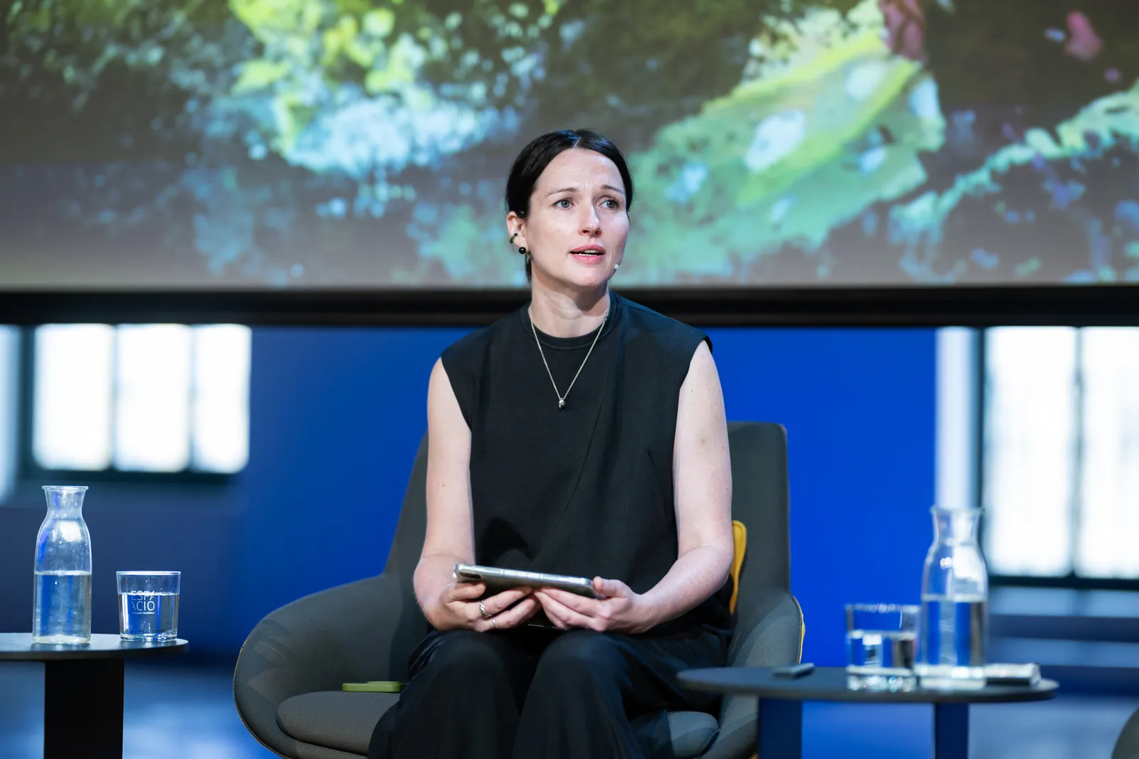 Woman in a sleeveless black top sits on a stage chair, holding a tablet during a panel discussion, with water carafes on side tables behind her.