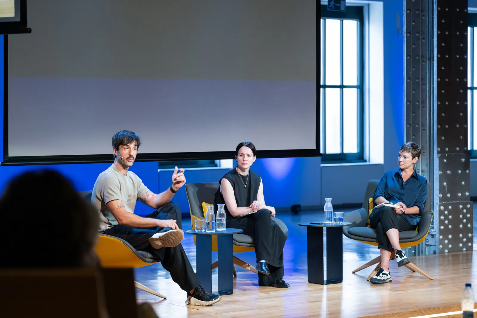 Three panelists sit in modern chairs on a stage: a man on the left speaks, a woman in the center listens, and a woman on the right takes notes; a large screen looms behind them and small tables hold water glasses.