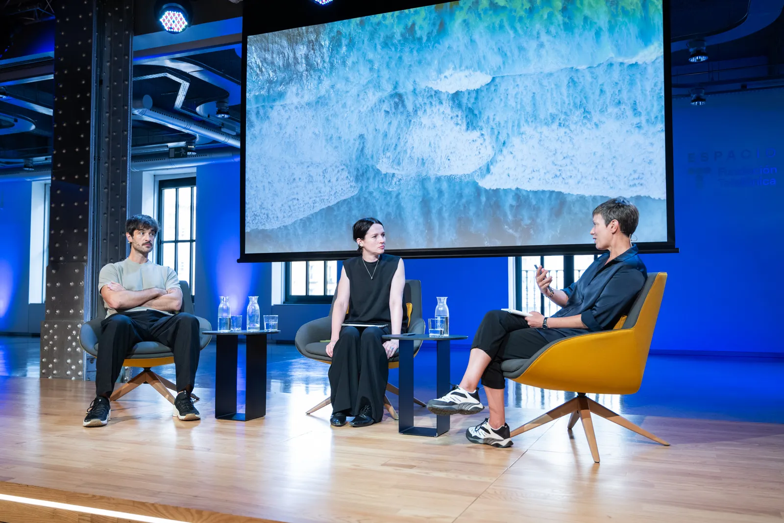 Panel discussion on a stage with three speakers seated in modern chairs, a large ocean-wave image on a screen behind them, and blue stage lighting with water pitchers on small tables.