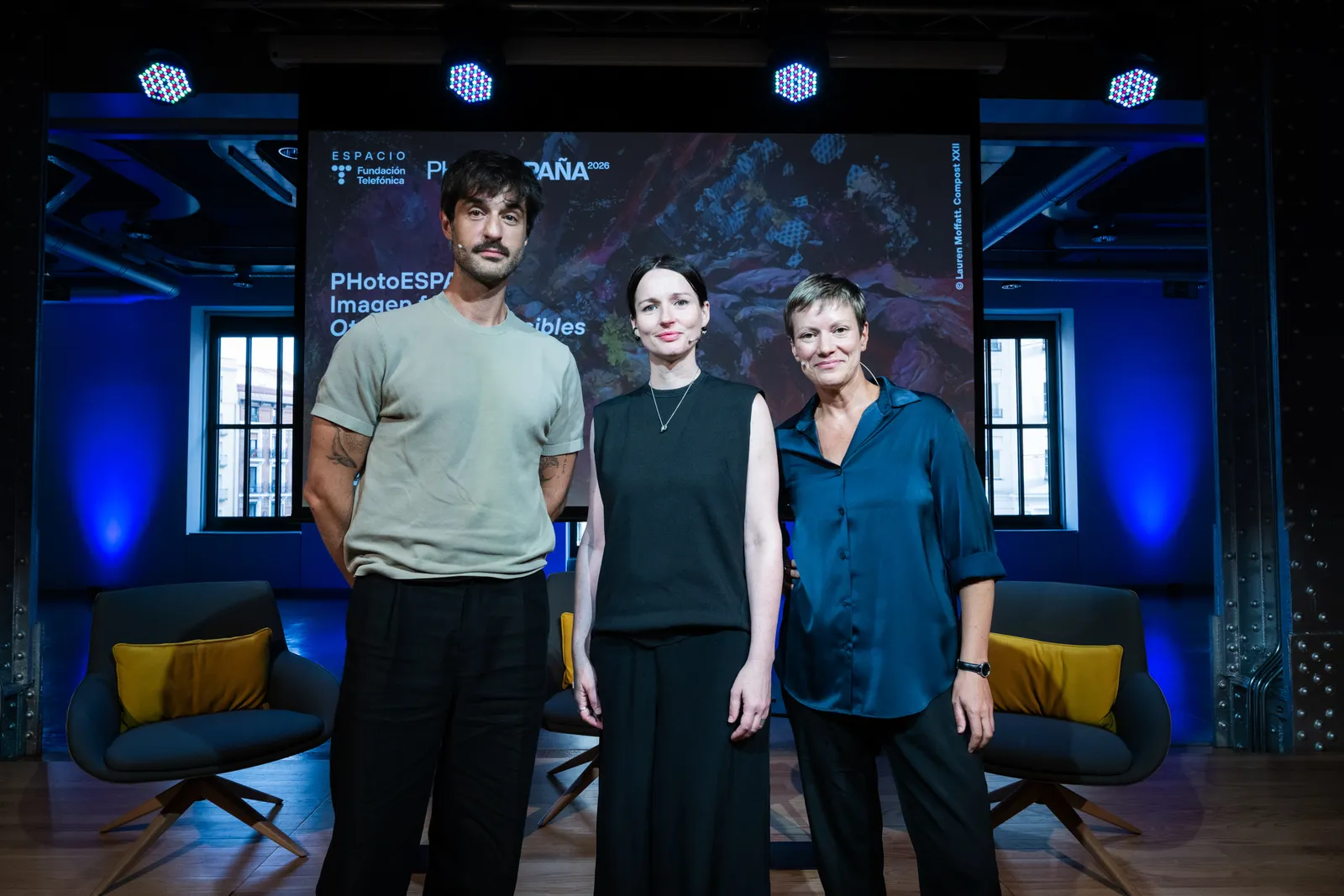 Three presenters pose on a dimly lit stage with a large screen behind them and blue lighting, seated chairs with yellow cushions visible to the sides.