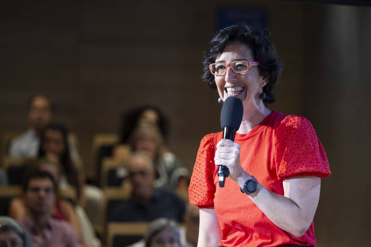 Woman with pink glasses and a bright red blouse smiles while speaking into a handheld microphone in front of an audience.
