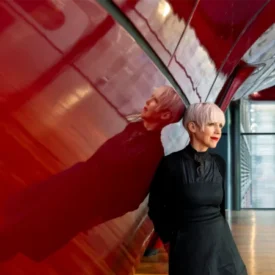 Woman with short blonde hair in a black dress and boots leans against a glossy red curved wall in a modern interior, her reflection visible on the surface.