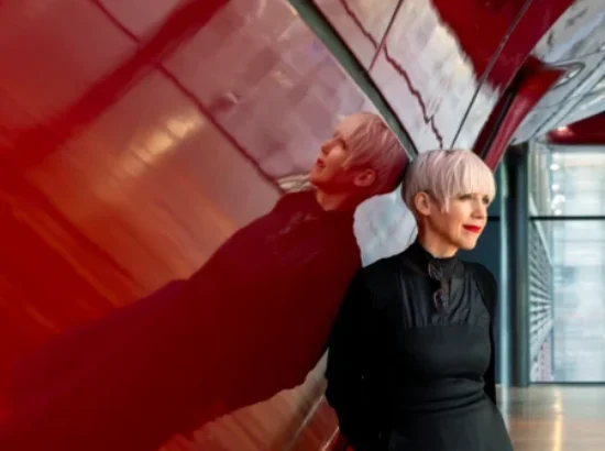Remedios Zafra with short blonde hair in a black dress and boots leans against a glossy red curved wall in a modern interior, her reflection visible on the surface.