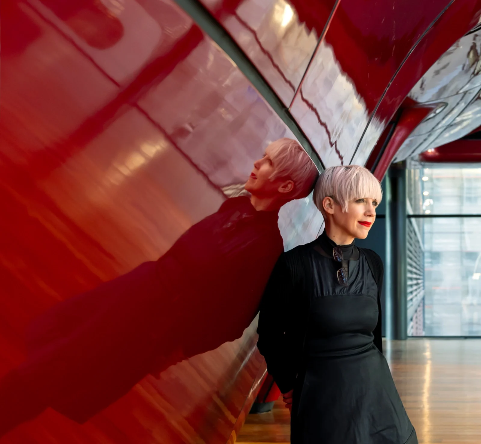 Woman with short blonde hair in a black dress and boots leans against a glossy red curved wall in a modern interior, her reflection visible on the surface.