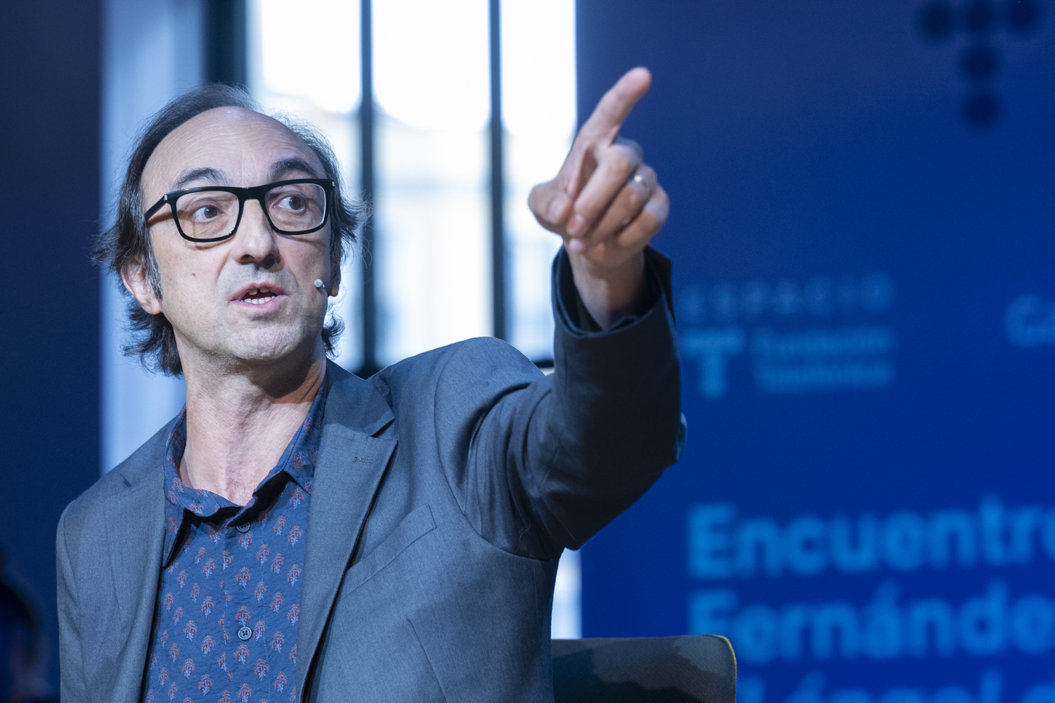 Middle-aged man with glasses and a blue patterned shirt gestures and points while speaking on stage.