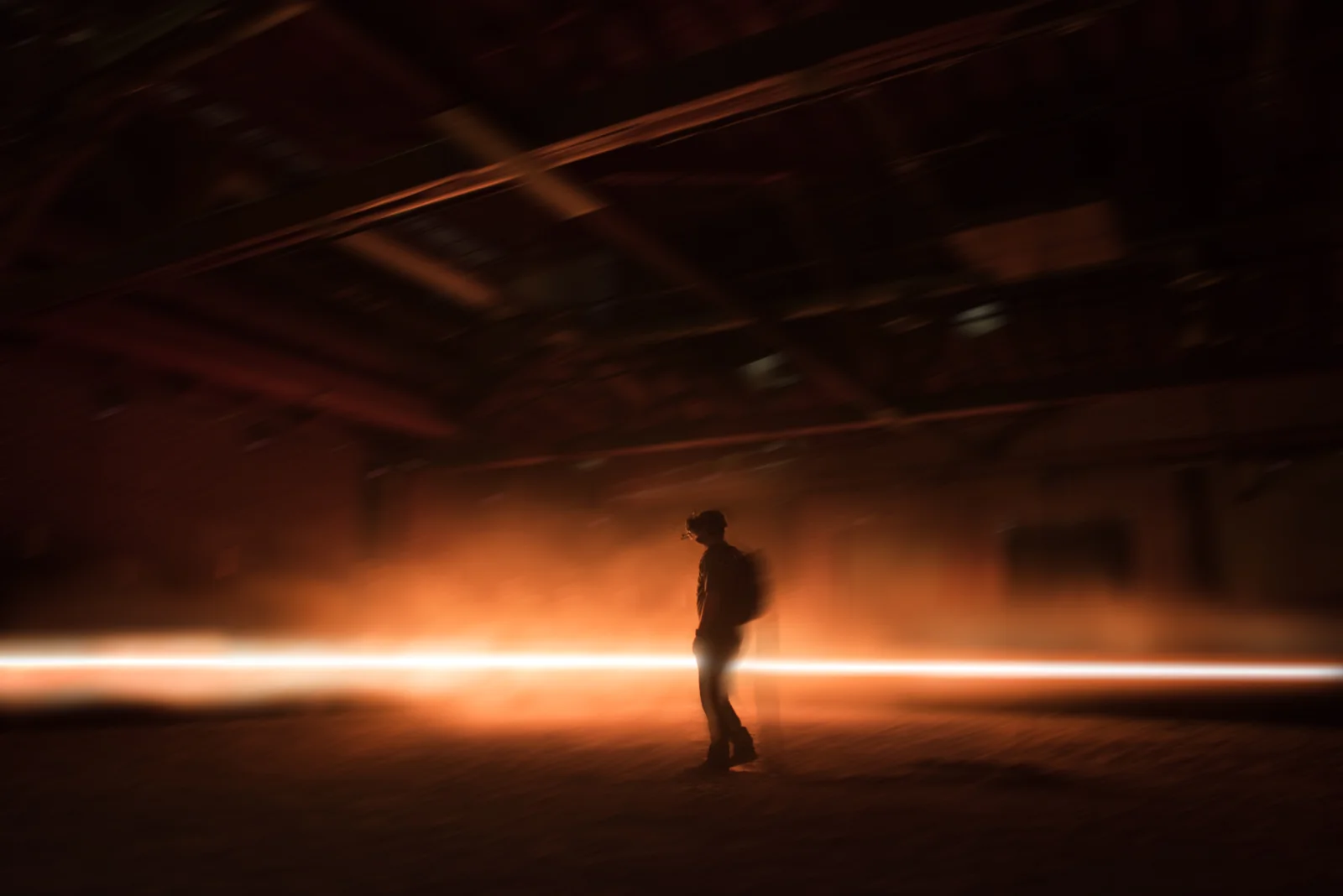 Una persona en silueta con mochila camina a través de un espacio industrial oscuro y humeante mientras una ráfaga de luz naranja pasa detrás de ella.