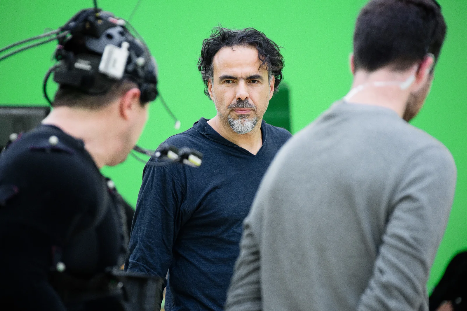 Alejandro G. Iñárritu, con barba canosa y camisa azul oscuro permanece de pie en un juego de rodaje, flanqueado por miembros del equipo frente a un fondo de pantalla verde.