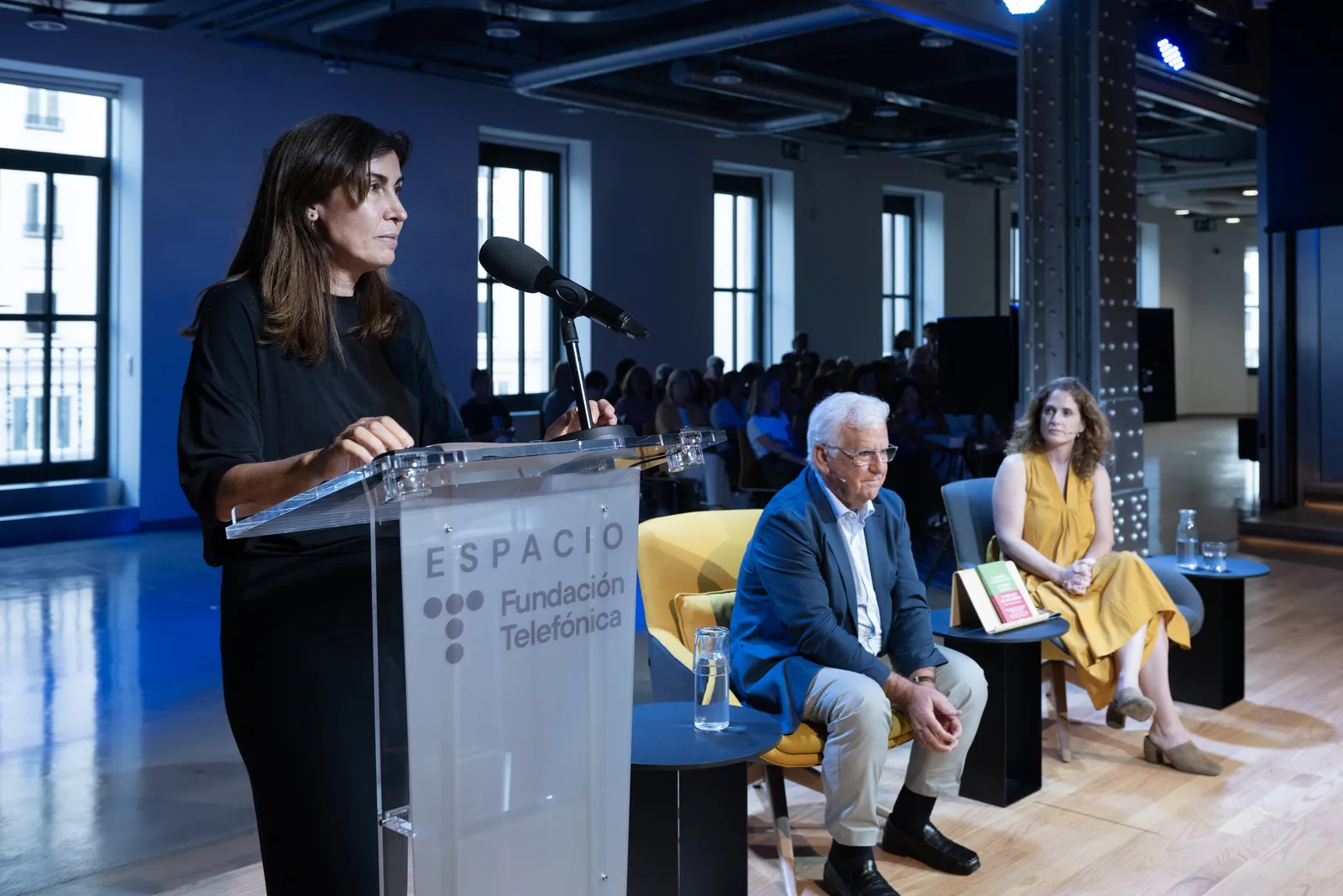 Woman in a black dress speaks at a podium labeled Espacio Fundación Telefónica, with two panelists seated behind her and an audience in the background.