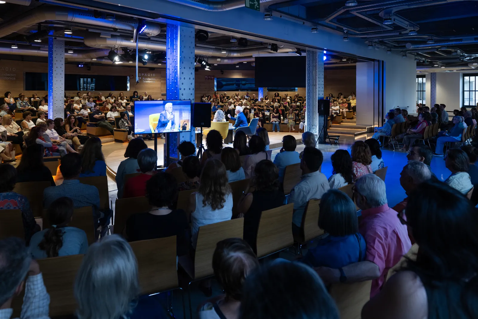 Panel discussion in a large auditorium with two speakers on stage and a screens displaying a speaker, surrounded by an attentive audience.