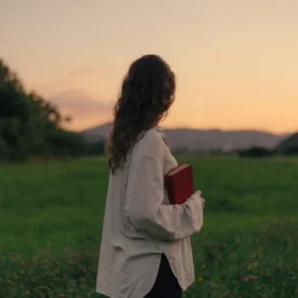 Person standing in a green field at sunset, holding a red book, seen from behind.